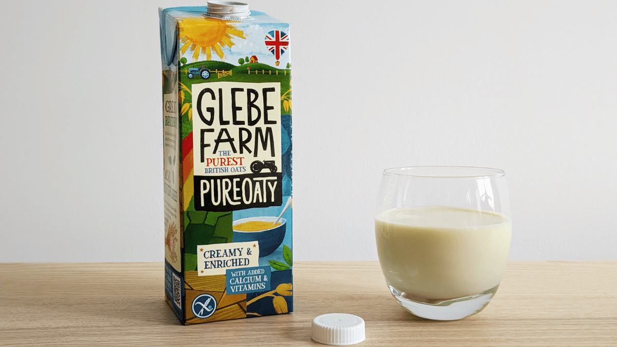 carton of oat milk beside a glass of poured oat milk on a wooden table top against a white background